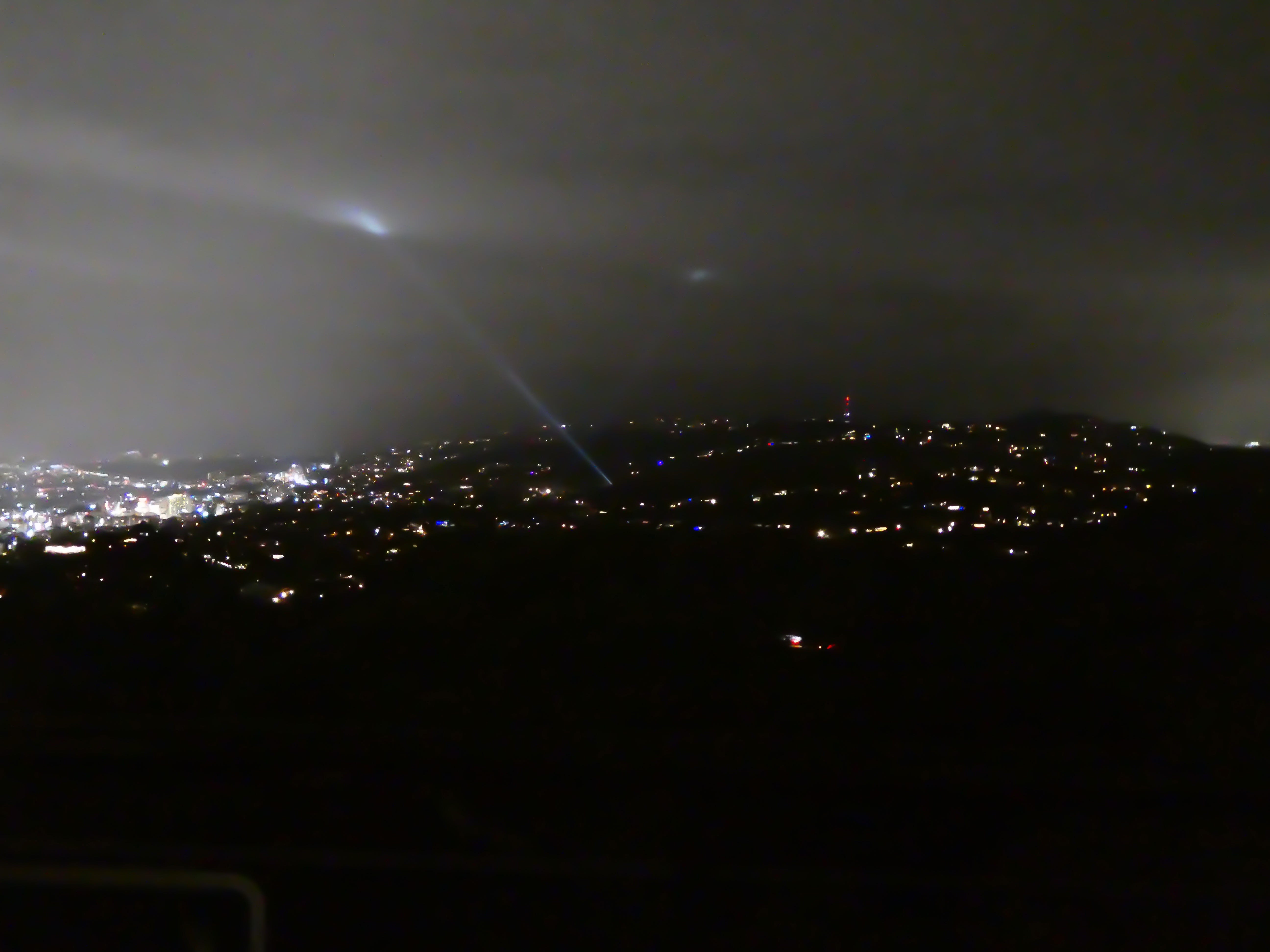 The Los Angeles landscape shrouded in dark and clouds. There are two lone search lights illuminating the darkness.